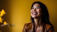 Young Asian woman in brown leather jacket laughing genuinely against yellow background, showing authentic joy and positive emotions in casual lifestyle portrait.
