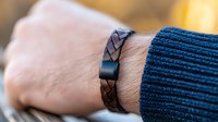Close-up of brown braided leather bracelet on male wrist wearing navy blue knit sweater, showing modern casual fashion accessory against blurred background.