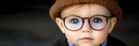 Adorable toddler boy with blue eyes wearing glasses and knit hat looking directly at camera with serious expression against dark background.