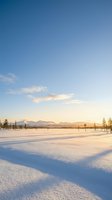Pristine snow-covered landscape with distant mountains under clear blue sky at golden hour. Peaceful winter scenery with sparse trees creates serene atmosphere.