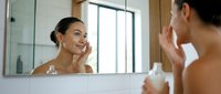 Young woman with olive skin tone applying skincare product to face while looking in bathroom mirror during morning beauty routine.