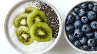 Fresh sliced kiwi and whole blueberries with chia seeds on white yogurt in ceramic bowls against white background, healthy breakfast concept.