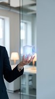 Person in dark business suit holding glowing holographic cube in modern office environment with glass partitions and natural light from windows.