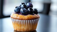 Fresh blueberry muffin topped with ripe berries and purple jam in white paper cup against blurred background, close-up view of homemade pastry.