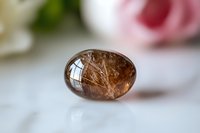 Polished rutilated quartz crystal egg with golden needle inclusions on soft white background, natural healing stone with rose petals bokeh effect.