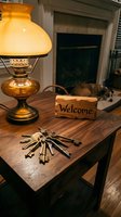 Cozy home entryway with vintage brass lamp, bundle of keys, and wooden welcome sign on dark wood table, fireplace in background.