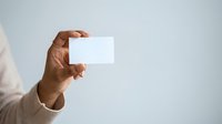 Man with dark skin tone wearing beige sleeve holding blank white business card against neutral background for branding and corporate identity mockup purposes.
