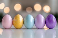 Colorful Easter eggs in pastel pink, gold glitter, blue, lavender and purple arranged in row with soft bokeh lights background and reflection on white surface.