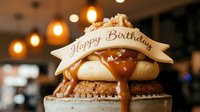 Gourmet birthday dessert with caramel drizzle and Happy Birthday banner topper, served in a cafe setting with warm bokeh lights.