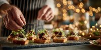 Chef garnishing gourmet appetizers on stone platter with microgreens, festive golden bokeh lights creating warm holiday atmosphere in background.