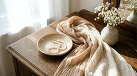 Elegant wooden table with cream fabric, ceramic bowl containing pearl jewelry, white flowers in vase creating luxurious home decor styling composition.