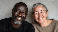 Mature diverse couple smiling together against gray wall, showcasing genuine happiness and connection in a warm, intimate portrait.