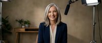 Mature woman with shoulder-length blonde hair, wearing dark blazer and cream blouse, smiling in professional recording studio with microphone and lighting equipment.