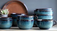 Handcrafted ceramic tea cups and bowls with turquoise reactive glaze and brown rim on neutral background. Artisanal pottery collection showcases organic textures.