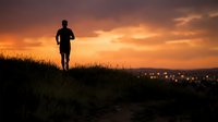Male runner jogging on hillside at sunset with city lights glowing in distance below, silhouetted against dramatic orange and golden sky, outdoor fitness and evening exercise.