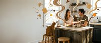 Young couple enjoying coffee together at modern kitchen counter with decorative geometric wall art and warm morning light creating cozy domestic atmosphere.