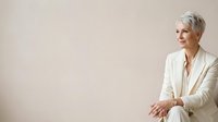 Mature woman with short white hair and fair complexion wearing cream blazer, seated against neutral background, professional business portrait.