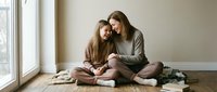 Mother and teenage daughter bonding together sitting on floor by window in cozy home interior wearing casual comfortable clothing during quality time.