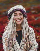 Young Caucasian woman with white dreadlocks and freckles wearing knitted winter hat and sweater smiling genuinely against autumn red foliage background outdoors.