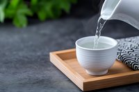 Pouring hot water from white ceramic teapot into cup on wooden tray. Traditional tea ceremony setup with striped napkin on dark surface for relaxation.
