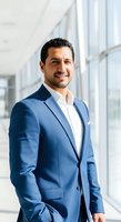 Portrait of a middle-aged male professional with dark hair and facial hair wearing a blue blazer and white shirt in a modern office corridor with natural light.