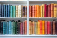 Colorful vintage books arranged by color spectrum on white wooden shelves, creating rainbow gradient effect in library or bookstore setting. Decorative hardcover spines display.