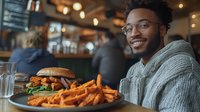Young African American man in glasses and gray sweater smiling at casual restaurant with gourmet burger and sweet potato fries on plate, warm ambient lighting creates cozy atmosphere.