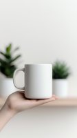 White ceramic mug held in feminine hand against minimal light background with soft green plants, mockup ready for custom design and branding projects.