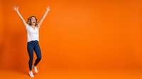 Excited young woman with red hair jumping with arms raised in celebration against vibrant orange background, expressing joy and success for lifestyle and motivation content.
