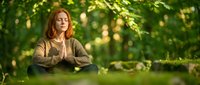 Middle-aged Caucasian woman practicing meditation in serene forest setting with dappled sunlight filtering through green foliage creating peaceful atmosphere.