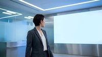 Professional Asian businesswoman in modern office looking at large blank presentation screen for corporate meetings and digital displays.