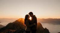 Romantic couple silhouette embracing at mountain summit during golden hour sunset with dramatic landscape and warm glowing light creating intimate moment.