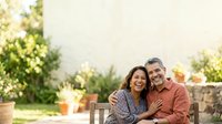Mature couple embracing on wooden bench in sunny garden setting with blooming flowers and green plants, enjoying outdoor leisure time together.