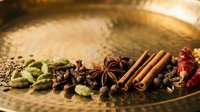 Assorted spices including cardamom, cloves, cinnamon sticks and red pepper arranged on a brass plate for cooking and culinary use.