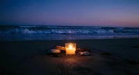 Glowing candle lantern on sandy beach at twilight with ocean waves and purple sunset sky creating peaceful romantic atmosphere for meditation and relaxation.