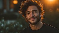 Happy young Middle Eastern man with curly hair smiling warmly in golden hour lighting. Casual portrait with bokeh background for lifestyle content.