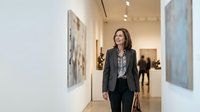 Mature woman with dark hair, Caucasian ethnicity, wearing professional business attire including charcoal blazer and patterned blouse, walking through contemporary art gallery with white walls and framed artwork, suitable for corporate, lifestyle, and cultural content.