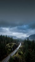 Aerial view of a winding mountain road cutting through dense evergreen forest with dramatic storm clouds overhead and distant peaks shrouded in mist, suitable for travel and nature themes.