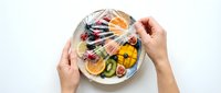 Hands holding plate with fresh sliced fruits including citrus, kiwi and berries wrapped in plastic film, food preservation and storage concept.