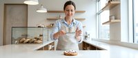 Middle-aged male baker with glasses wearing apron stands confidently at counter with fresh bread and pastries in modern bakery kitchen with natural light.