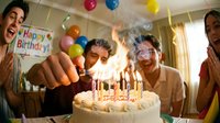 Friends lighting candles on a white birthday cake at a festive party with colorful balloons and a Happy Birthday banner in the background.