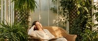 Young woman relaxing on wicker chair in sunlit greenhouse surrounded by lush green plants and natural light for wellness and tranquility concepts.