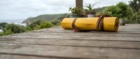 Yellow yoga mat rolled up with leather strap on wooden deck overlooking green hills and ocean, outdoor fitness and wellness retreat setting for mindful exercise.