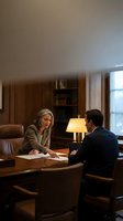 Two professionals in business attire conduct a meeting at a wooden desk in a traditional office setting with warm lighting, bookshelves, and formal furnishings for corporate communications.
