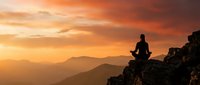 Person meditating on mountain peak at sunset with orange sky and layered mountain ranges creating peaceful spiritual atmosphere for wellness content.