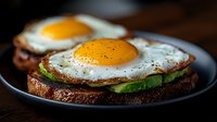 Sunny side up eggs on dark rye toast with fresh avocado slices and cracked black pepper, served on black plate against rustic wooden background.