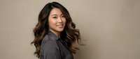 Young Asian businesswoman with long wavy hair smiling confidently in professional gray blazer against neutral beige background for corporate headshot.
