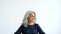 Senior woman with long gray hair wearing dark navy blue shirt, smiling while looking to the side against white background for portrait and lifestyle use.