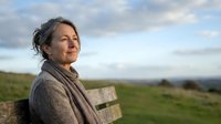 Mature woman enjoying peaceful moment outdoors on wooden bench in countryside landscape with cloudy sky background for wellness and retirement concepts.