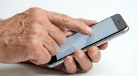 Senior adult male with aged, wrinkled hands holding and using a smartphone on white background, representing technology adoption among older generations.